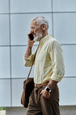 A mature gay man with a beard and a tattoo on his arm walks past a modern building while talking on his phone.