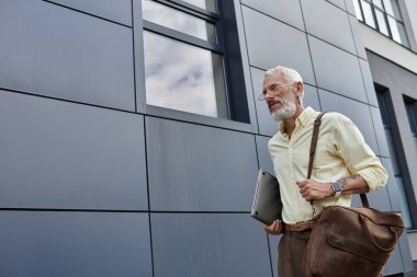 Sakallı bir adam, elinde bir laptop ve kahverengi bir çantayla modern bir binanın önünde yürüyor..