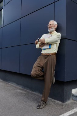 A mature gay man with a beard leans against a modern building, smiling as he daydreams. clipart