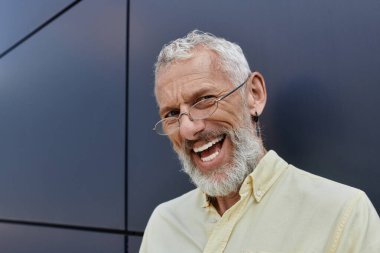 Sakallı, orta yaşlı bir adam yürekten gülüyor. Parlak gülüşü, modern bir binanın arka planında güneş tarafından aydınlatılıyor..