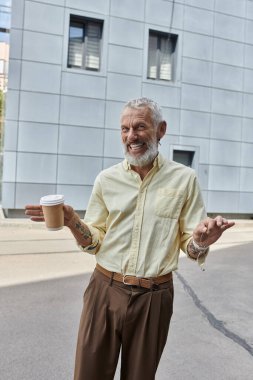 Sarı gömlekli bir adam modern bir binanın önünde elinde bir fincan kahve ve gülümsüyor..