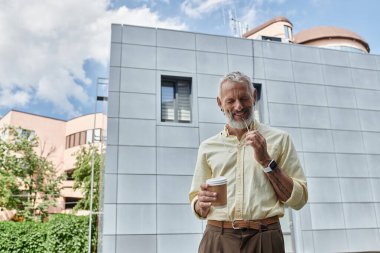 A smiling middle-aged man with a beard enjoys a coffee break outside a modern building. clipart