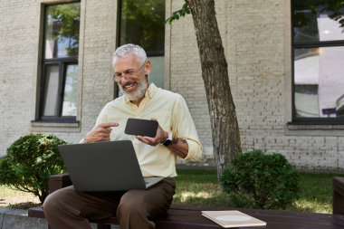 Sakallı, olgun bir eşcinsel, modern bir binanın önündeki bankta oturuyor. Telefonunu ve laptopunu kullanarak gülüyor..