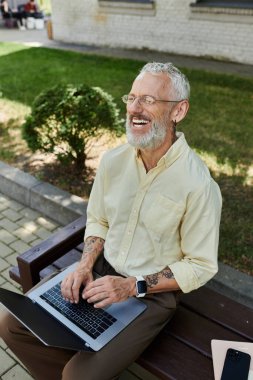Sakallı bir adam binanın yanındaki bankta oturuyor, dizüstü bilgisayarda yazıyor ve gülüyor..
