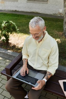 Sakallı, olgun bir adam dışarıda bir bankta dizüstü bilgisayar kullanarak oturuyor..