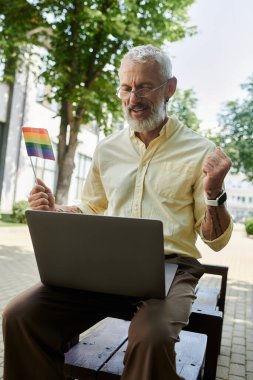 A gay man celebrates with a rainbow flag while working on his laptop outdoors. clipart