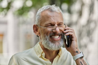 Sakallı ve gözlüklü bir adam dışarıda telefonla konuşurken gülümsüyor..