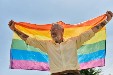 A smiling, bearded man holds a rainbow flag high in the air, celebrating pride and acceptance. clipart