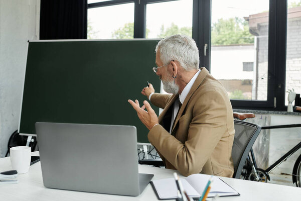 A middle-aged man with a beard teaches online, gesturing towards his laptop with a blackboard behind him, lgbtq teacher