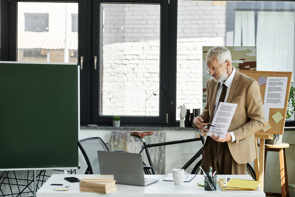 A bearded man in a suit and glasses stands by a laptop, holding a paper and teaching a lesson online