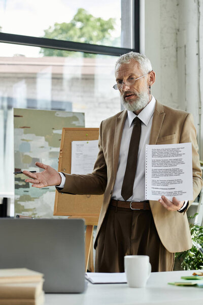 A middle-aged, bearded teacher wearing a suit, glasses, and a tie stands in front of a laptop, leading an online class.