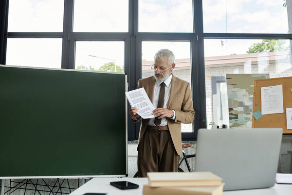 A middle-aged, bearded teacher in a suit looks over notes in a classroom before going online to teach, lgbtq teacher