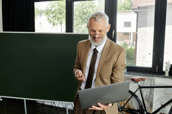 A middle-aged man with a beard, wearing a suit and tie, smiles while holding a laptop and teaching an online lesson.