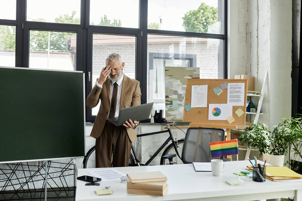 A mature LGBTQ teacher, with a beard, teaches a class online using a laptop in his modern classroom.
