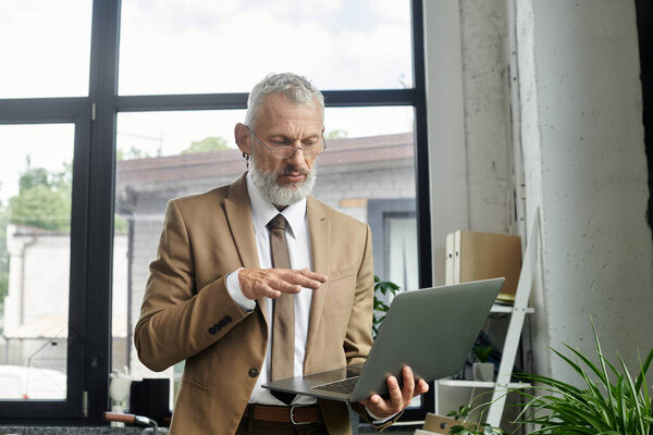 A mature LGBTQ teacher with a beard conducts an online lesson, standing in a modern office with a window in the background.