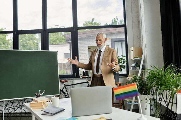 A middle-aged gay man with a beard teaches an online lesson in front of a laptop and a whiteboard, a rainbow flag is visible.