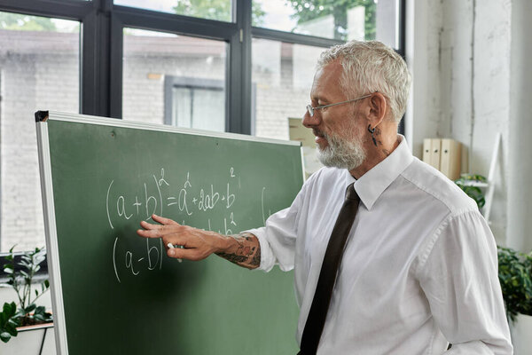 A middle-aged man with a beard teaches an online math class, pointing to a formula on a chalkboard.