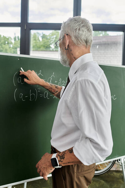 Middle-aged teacher with a beard, glasses, white shirt writes an equation on a chalkboard while teaching online in his classroom