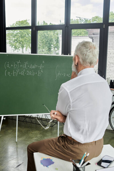 A mature LGBTQ teacher with a beard sits by a chalkboard during an online lecture.