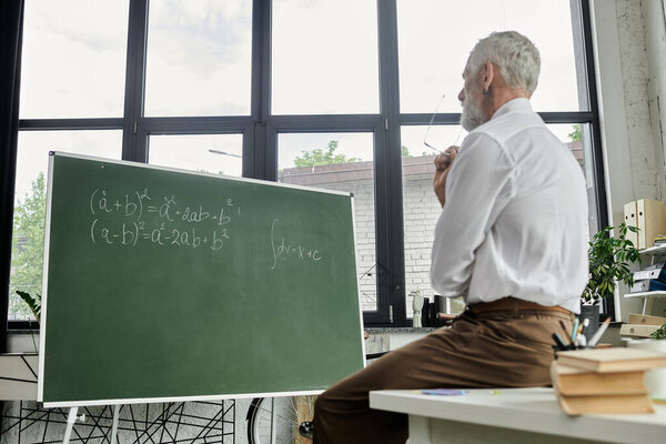 A mature teacher with a beard sits by a whiteboard with math equations, teaching online through a laptop.