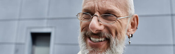 A middle-aged man with a beard and glasses smiles broadly, standing near a modern building.
