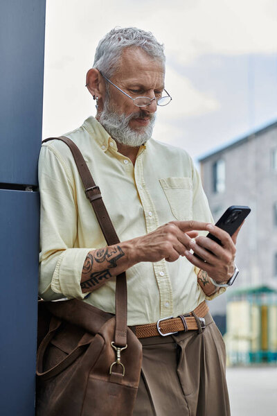 A mature gay man with a beard checks his phone, leaning against a building in a modern urban setting.