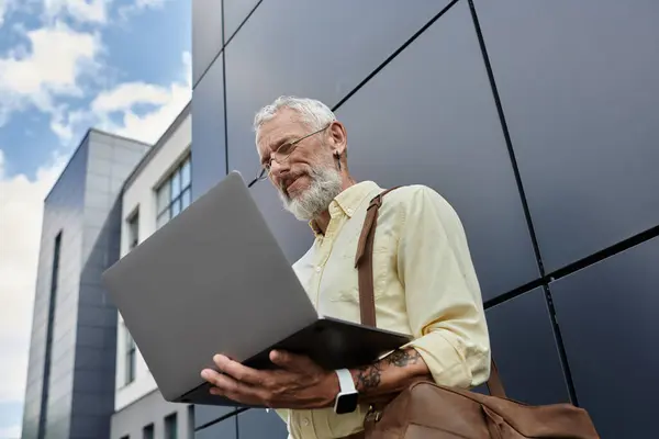 stock image A mature gay man with a beard checks his laptop while standing outside a modern building.