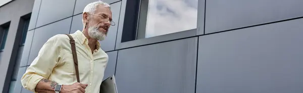 stock image A mature gay man with a beard and glasses walks by a modern building, carrying a bag.