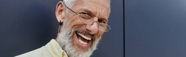 A bearded man with glasses laughs outdoors in front of a modern building.