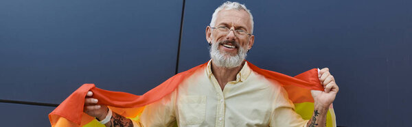 A middle-aged man with a beard smiles while holding a rainbow flag in front of a modern building.