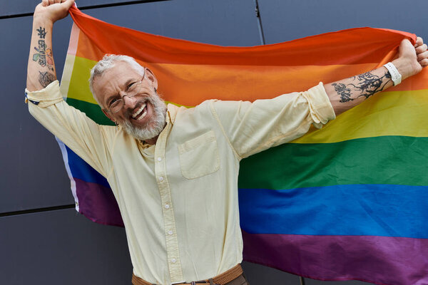 Мужчина средних лет с бородой широко улыбается, держа радужный флаг гордости перед современным зданием.