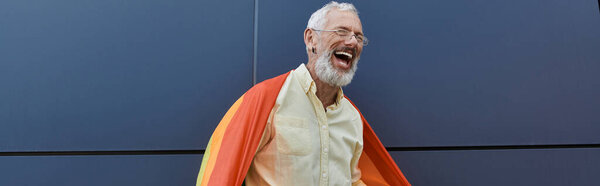 A mature man with a beard laughs outside a modern building, wearing a pride flag like a cape.