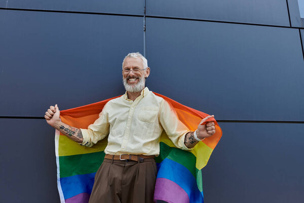 Мужчина с бородой широко улыбается, держа радужный флаг перед современным зданием.