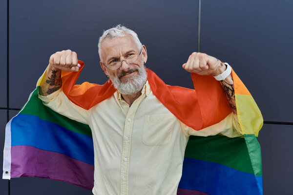 Улыбающийся, зрелый мужчина с бородой гордо стоит с радужным флагом на плечах..