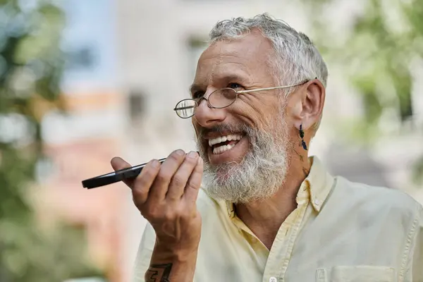 stock image A joyful man, sporting a beard and glasses, enjoys a sunny afternoon outdoors, laughing while using his phone.