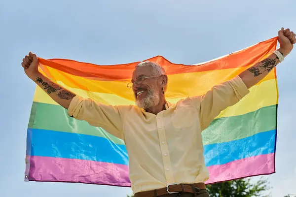 stock image A smiling, bearded man holds a rainbow flag high in the air, celebrating pride and acceptance.