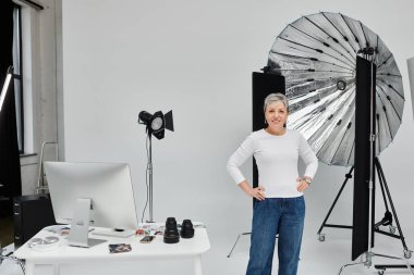 Beyaz gömlek ve kot pantolon giymiş olgun bir kadın, bir fotoğraf stüdyosunda güvenle duruyor..