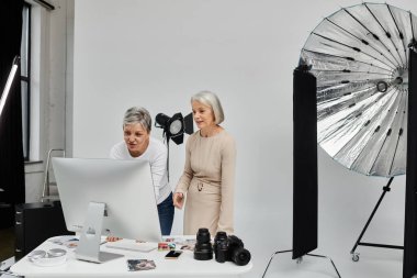 İki olgun kadın, bir fotoğrafçı ve bir model, bir fotoğraf stüdyosundaki bilgisayar ekranındaki görüntüleri gözden geçirirler..