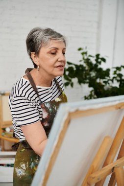 A mature woman in an art studio, wearing a striped shirt and an apron, looks at a blank canvas on an easel. clipart