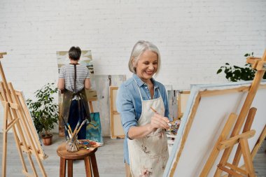 Two mature women are painting in an art studio. One woman is smiling as she holds a paintbrush and palette. clipart