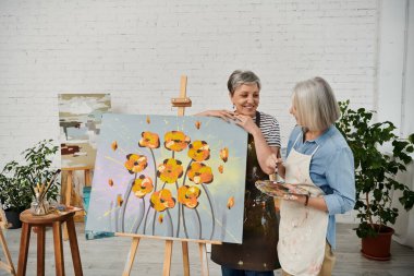 Two women, a mature lesbian couple, paint together in an art studio, sharing a smile and a moment of creativity. clipart