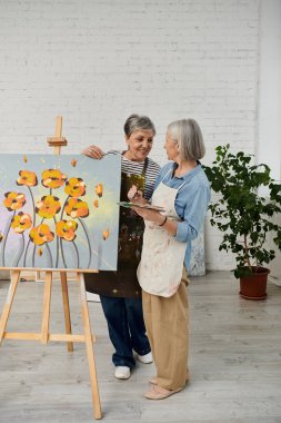 İki kadın bir sanat stüdyosunda durur, biri sehpada tuvali boyarken diğeri paleti tutar ve gözlemler..