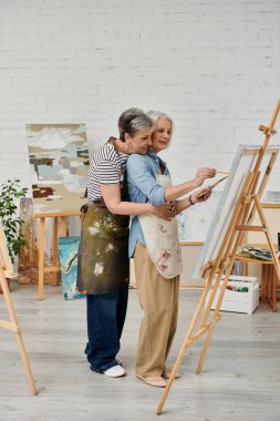 A couple in an art studio, one woman painting while the other watches, arms around her waist. clipart