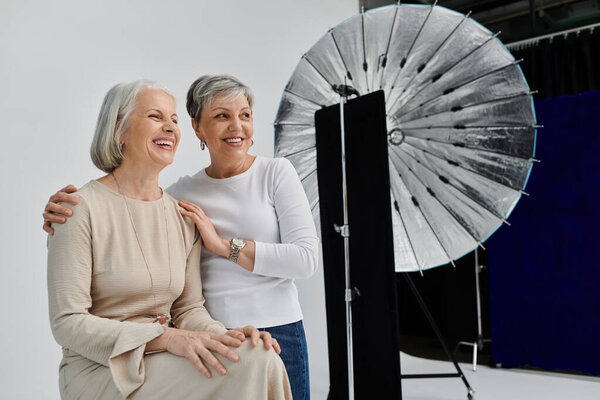 Two mature women, dressed in casual clothes, smile and embrace during a photoshoot in a studio.