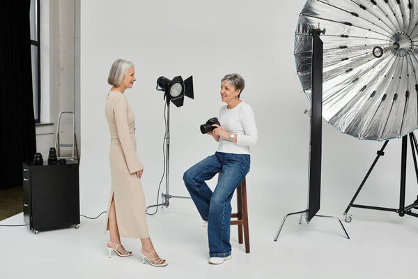 Two women, one holding a camera, the other standing in front of a backdrop, enjoying a photoshoot in a studio.