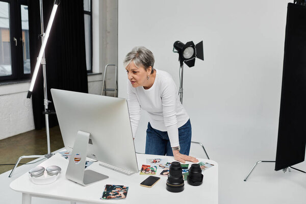 Женщина наклоняется над компьютером в фотостудии, рассматривая изображения с серьезным выражением.