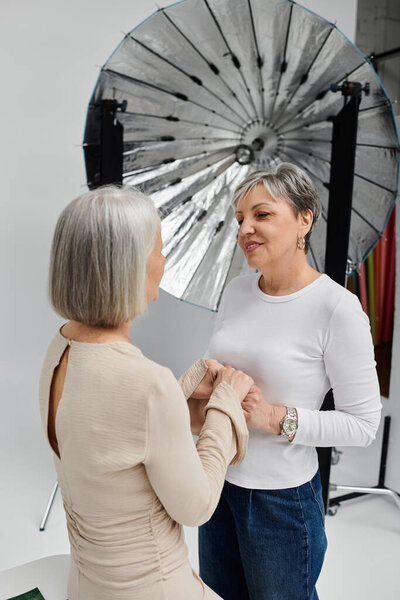 A lesbian couple in a studio, one taking photos, the other modeling. They hold hands and share a loving glance