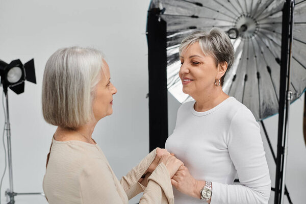 A mature lesbian couple, one a photographer and the other a model, share a tender moment in a studio setting.