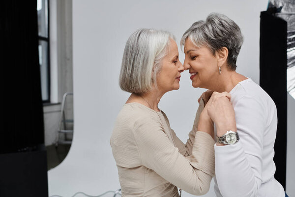 Two mature women stand close together in a studio, smiling and touching noses.