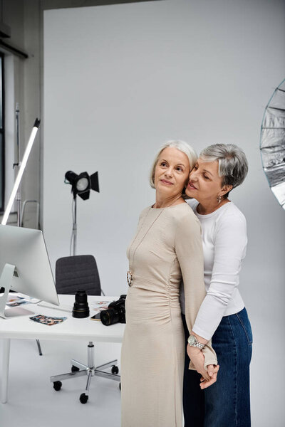 Two mature women, a photographer and her model, stand close in a photo studio, holding hands.
